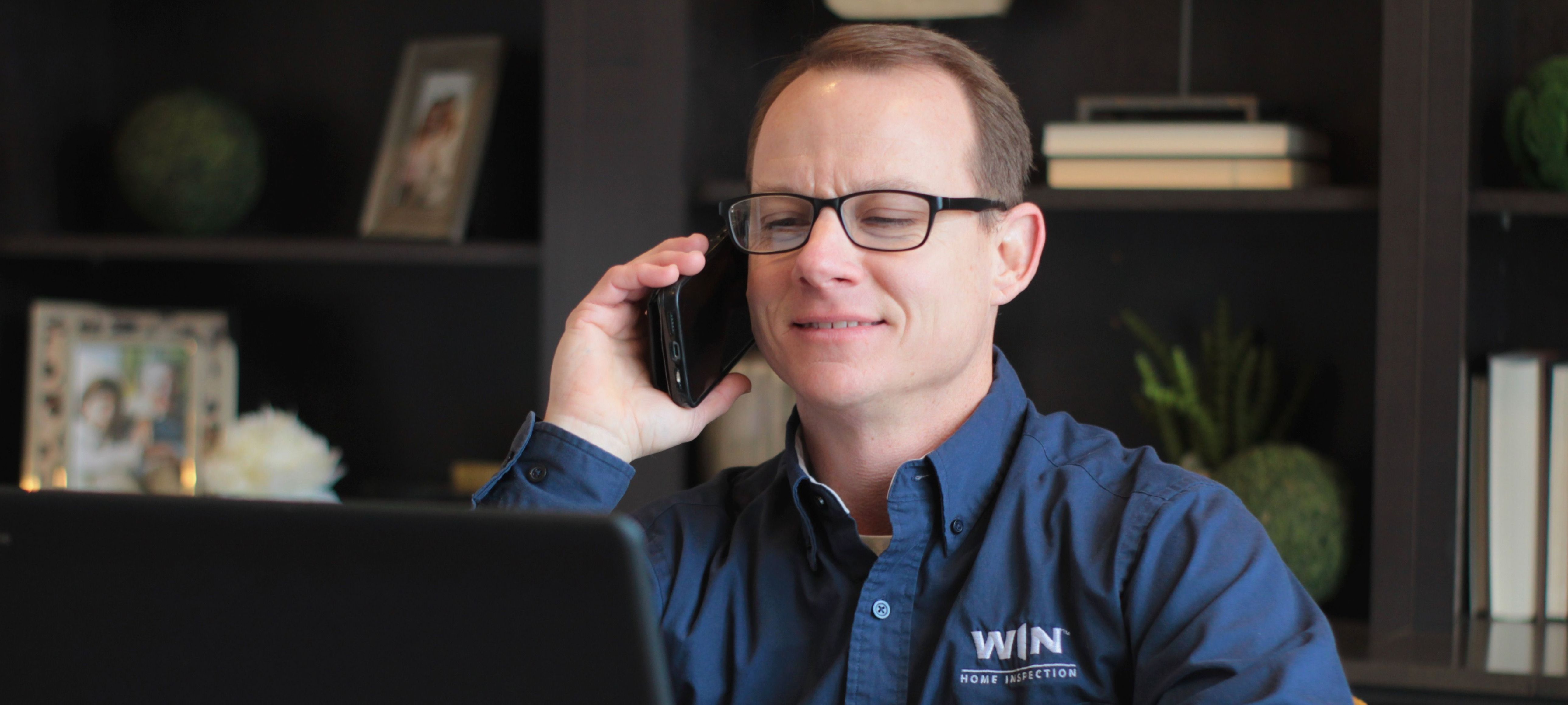 WIN Home Inspector holding a smartphone to their ear while seated at a desk with laptop.